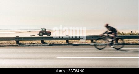 Pacific Coast Highway ciclista professionista e un trattore rosso che passa nella direzione opposta sulla sabbia di fronte all'Oceano Pacifico. Foto Stock