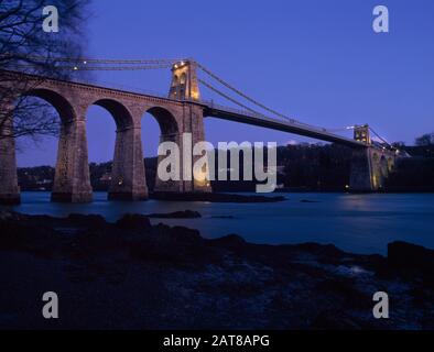 Telford'S Bridge, Menai Suspension Bridge, Menai Bridge, Anglesey, Galles Del Nord. Guardando dal litorale su Anglesey fino al ponte illuminato. Foto Stock