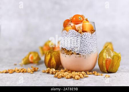 Il bicchiere di Smoothiein a strati sani con chia vede il pudding, mescolato con cereali e condito con persimmon arancione e frutti di physalis Foto Stock