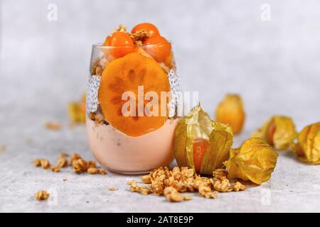 Il bicchiere di Smoothiein a strati sani con chia vede il pudding, mescolato con cereali e condito con persimmon arancione e frutti di physalis Foto Stock