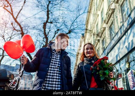 San Valentino amore coppia giorno. Fidanzato e fidanzata a piedi con fiori di rose e palloncini a forma di cuore in città Foto Stock