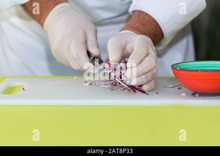 Lo chef maschile raccoglie i dadi di cipolla rossa appena tagliati da un tagliere con un coltello. Concetto di cucina. Foto Stock