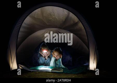 Ragazze di pre-teen che posano in una tenda del campeggio avvolta in una borsa del sonno che legge un libro con la torcia di notte durante le feste estive Foto Stock