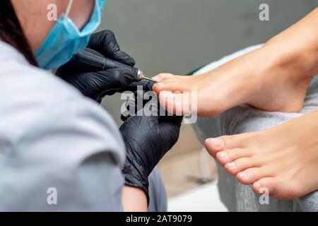 Pedicurist le mani in guanti con attrezzo speciale facendo pedicure femminile di chiodi sui piedi. Trattamento piedi in spa salone, vicino. Foto Stock