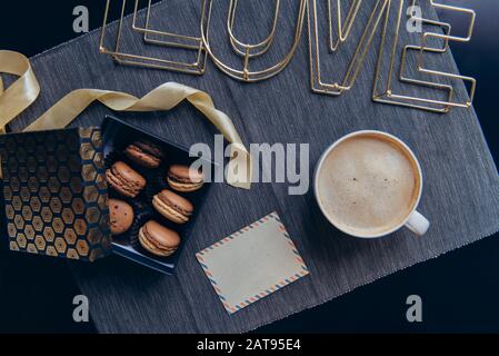 Colazione Accogliente con vista dall'alto per gli amanti. Mattinata romantica in stile nordico noir. Tazza da caffè, scatola con macaron, cartolina retrò vuota e filo parola Love on g. Foto Stock