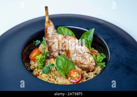 Gamba di coniglio fritto con riso su una piastra rotonda nera. La decorazione è decorata con verdure e pomodori. Sfondo bianco Foto Stock
