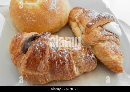 Due croissant italiani freschi ripieni di vaniglia e creme al cioccolato, accompagnati dalla brioche rotonda con toppo, su un piatto bianco con tovaglioli. Foto Stock