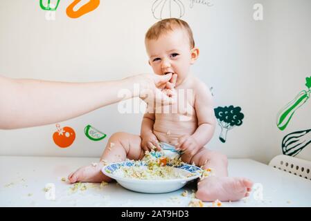 Sporco e sorridente bambino su un tavolo bianco essendo alimentato da sua madre la mano, mentre ridono mentre provando il metodo blw. Foto Stock