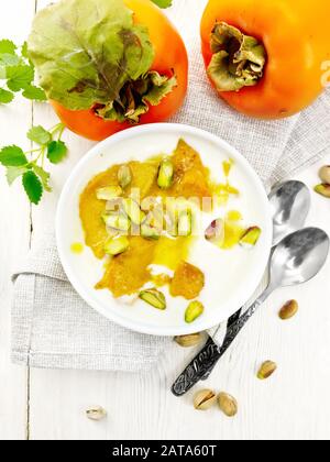 Dessert di yogurt, persimmon e miele con vaniglia, cardamomo e pistacchi in una ciotola su un tovagliolo, menta e cucchiaio sullo sfondo di legno chiaro b Foto Stock