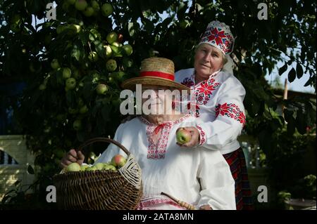 Coppia slava anziana in camicie ricamate. Nonno e donna in costumi nazionali con un cesto di mele. Pensionati bielorussi con un raccolto. Foto Stock