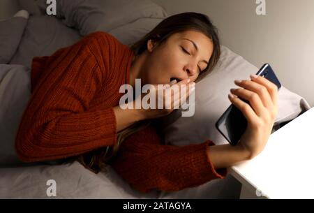 Bella giovane donna sdraiata a letto e tendente guardando i social media su smartphone. Ragazza stanca guardando mobile o ascoltando musica e sonnolenza, femmina l Foto Stock