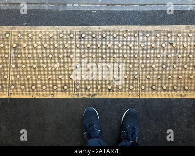 Attenzione al divario: In attesa sulla piattaforma di una stazione della metropolitana di Londra. POV, guardando giù la linea di divisione goffrata, il pavimento e le scarpe Foto Stock