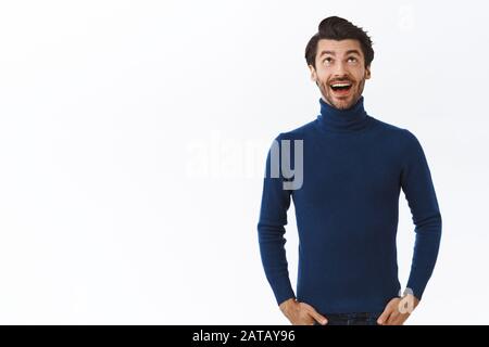 Un uomo d'affari entusiasta e fortunato, con un maglione blu a collo alto, che guarda in alto e sorridente divertito, tiene le mani in tasca, divertito come qualcosa di buono Foto Stock