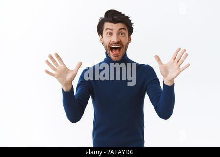 Un uomo elegante e dall'aspetto accattivante, con barba, capelli scuri, un pullover ad alto collo, le mani alzate che si scuotono per trionfare, celebrando le eccellenti novità, smilin Foto Stock