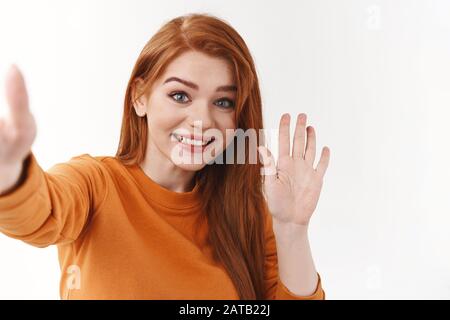 Donna simpatica e in uscita in una maglione arancione, porta il braccio con lo smartphone o la fotocamera, fai un selfie, fai un'onda con il ciao o ciao Foto Stock