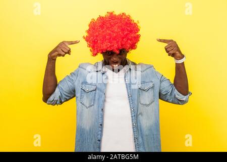 Ritratto di eccitato divertente spensierato uomo indossando camicia denim che punta alla parrucca rossa curly sulla sua testa e sorridente felice, stile di vita ottimista facile. Indoo Foto Stock