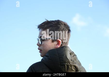 Un giovane ragazzo di 12 in occhiali con un viso fangoso che guarda al lato con sfondo blu cielo Foto Stock