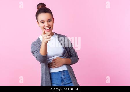 Ehi, tu! Ritratto di bruna gioiosa ragazza adolescente con acconciatura bun in abiti casual che punta alla macchina fotografica che tiene il suo stomaco e ridendo fuori forte, h. Foto Stock