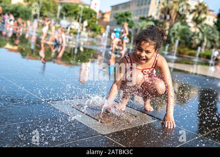 Il bambino si diverte con uno dei jet d’acqua che si trovano nel Mirroir d’eau sulla nuova piazza urbana, la Promenade du Paiilon a Nizza, Francia Foto Stock