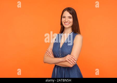 Attraente donna brunetta in denim abito in piedi mezzo girato con le mani incrociate e sorridente con denti bianchi, essere sano felice soddisfatto, espressi Foto Stock