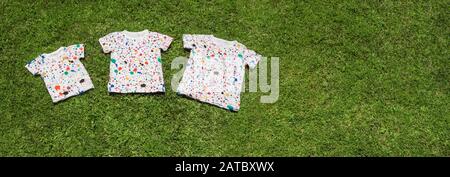 T-shirt sdraiata su erba verde. Campo estivo per bambini, concettuale. Grande sfondo familiare. Magliette colorate su sfondo verde erba. Banner. Vista dall'alto. Spazio di copia Foto Stock
