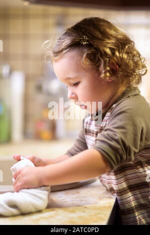 Сute bambina di due anni lava i piatti in cucina. Il bambino aiuta i genitori. Il bambino piccolo piacevole impara la famiglia. Toddler pulisce le sue mani con un a. Foto Stock