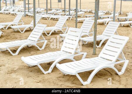Sedie da spiaggia in bassa stagione. Molti lettini liberi su una spiaggia di sabbia. Panorama di molti lettini in plastica bianca sulla riva del mare. Deserta resort bea Foto Stock