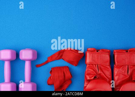 paio di guanti da boxe in pelle rossa, fascia elastica in tessuto rosso per le mani, manubri in plastica lilac su sfondo blu, vista dall'alto, copia spazio Foto Stock