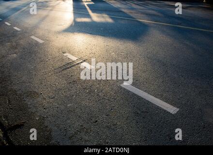 La luce del mattino cade su una superficie di asfalto vuota, con ombre, segnaletica stradale bianca e gialla e irregolarità di superficie di interesse. Foto Stock