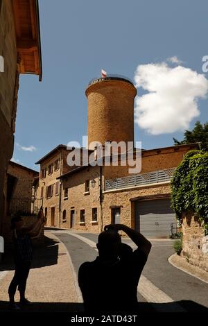 Un uomo dalla silhouette fotografa la Torre Oingt in una luminosa giornata di sole, mentre una nuvola bianca scivola attraverso il cielo blu dietro le pietre dorate dei villaggi Foto Stock