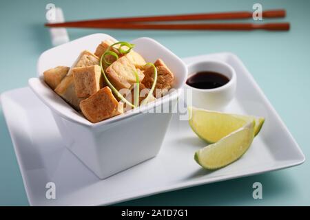 Tofu fritto servito con salsa di soia, fette di lime e cipolla verde su un piatto bianco di primo piano Foto Stock