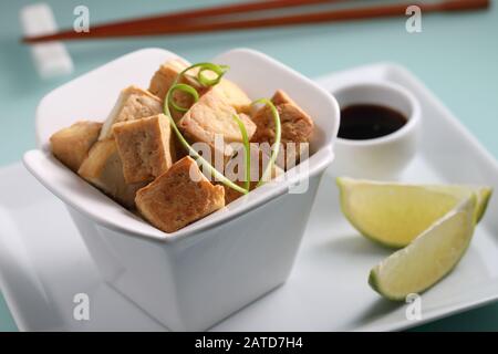Tofu fritto servito con salsa di soia, fette di lime e cipolla verde su un piatto bianco di primo piano Foto Stock