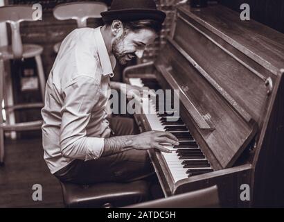 Alla moda uomo con elegante Cappello e barba cercando la riproduzione di vintage in legno antico pianoforte - Giovane artista nel cocktail bar - bianco e nero - editing Fo Foto Stock