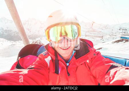 Felice sciatore scattare foto selfie con fotocamera smartphone seduta su skilift - giovane uomo divertirsi in inverno vacanza neve resort con luce posteriore - Spo Foto Stock
