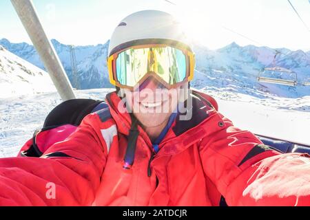 Felice sciatore tenendo selfie foto con smart cellulare fotocamera seduto su sci lift - Giovane avendo divertimento in inverno neve località vacanza con retro illuminazione Foto Stock