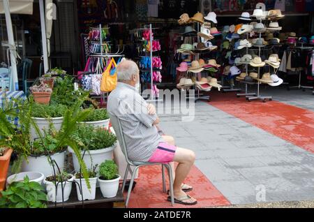 Sarti, Greece, June 16, 2018: Old street vendor sitting in front of store selling hats and slippers in touristic place Sarti, Sithonia Chalkidiki, Gre Foto Stock