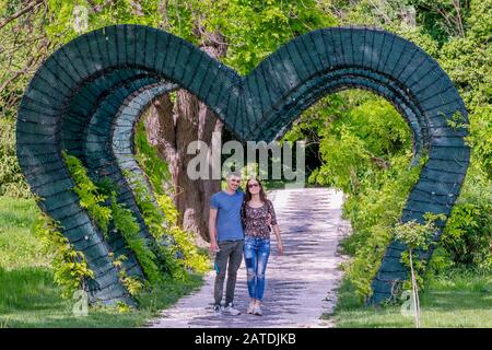 Giovane coppia impegnata camminare sotto un arco verde a forma di cuore nel parco pubblico, Craiova, Romania Foto Stock