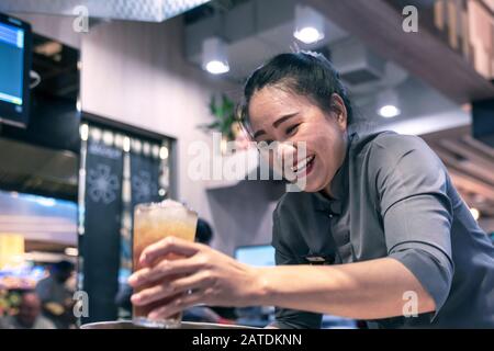 Thailandia, Phuket, Patong. 3 gennaio 2020: Una cameriera ridente serve a un cliente un bicchiere di Cola con ghiaccio. Atmosfera piacevole, servizio eccellente. Foto Stock