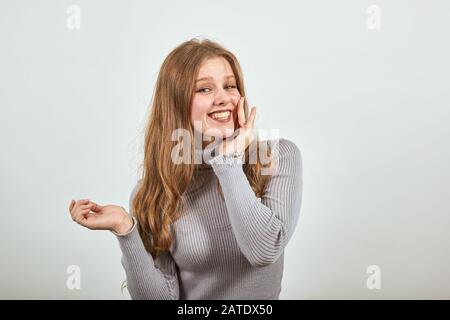 giovane bella donna dai capelli rossi in maglione grigio tiene la mano vicino al suo viso Foto Stock