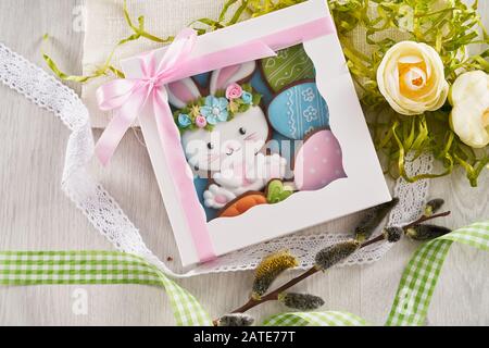 Primo piano di biscotti glassati a forma di bunny bello in wreath, carota e tre uova in scatola regalo, ramo salice, fiori secchi e nastri isolati su sfondo bianco. Dolce fatto in casa. Foto Stock