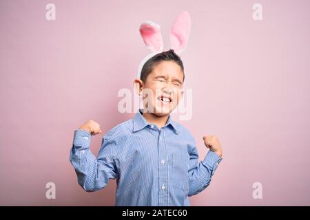 Giovane ragazzino che indossa orecchie di coniglio di pasqua su sfondo rosa isolato molto felice ed eccitato facendo il gesto del vincitore con le braccia sollevate, sorridenti e. Foto Stock