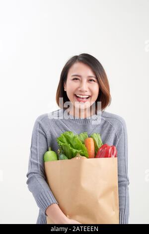 Bella giovane donna in grembiule tenendo carta shopping sacchetto pieno di verdure fresche e sorridente Foto Stock