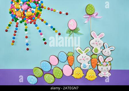 Vista dall'alto di colorati biscotti glassati allo zenzero e palline di cioccolato isolate su sfondo blu e viola. Deliziosa pasticceria fatta in casa a forma di animali di pasqua, uova, sole e carote. Foto Stock