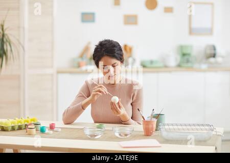 Ritratto di donna adulta moderna pittura uova di Pasqua mentre seduto al tavolo in accogliente cucina interno, copia spazio Foto Stock