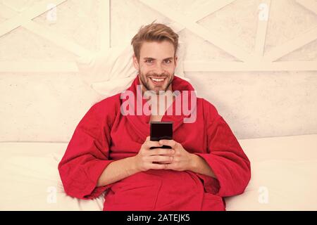 Vivere lo stile di vita mobile. Allegro guy utilizzando il dispositivo mobile a letto. Bel uomo sorridente con il cellulare in mano. La comunicazione mobile e tecnologie wireless in casa. Foto Stock