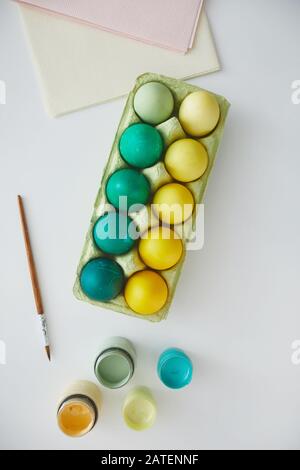 Vista dall'alto delle uova di Pasqua dipinte di verde e giallo in cassa disposte in composizione minima con pennello su sfondo bianco, spazio di copia Foto Stock