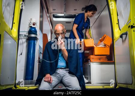 L'uomo respira attraverso la maschera di ossigeno, giovane infermiera in uniforme medica apre il sacchetto medico nell'automobile dell'ambulanza Foto Stock