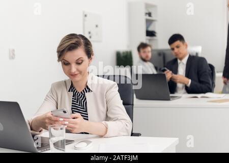 Giovane donna guarda il suo telefono sorprendentemente durante la giornata lavorativa in ufficio, i suoi colleghi siedono un tavolo accanto a lei Foto Stock