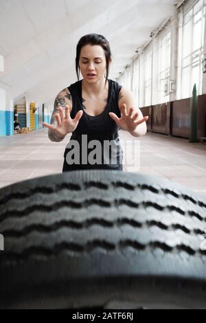 Giovane atleta di sesso femminile caucasica tatuata con capelli scuri che lanciano pneumatici pesanti Foto Stock