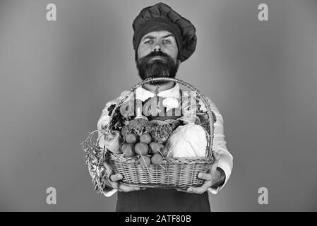 Lo chef tiene cavolo, ravanello, broccoli con lattuga, pepe e aglio. Uomo con barba su sfondo blu. Concetto di cucina sana. Cucinare con il viso stretto in uniforme borgogna tiene le verdure nel cesto Foto Stock